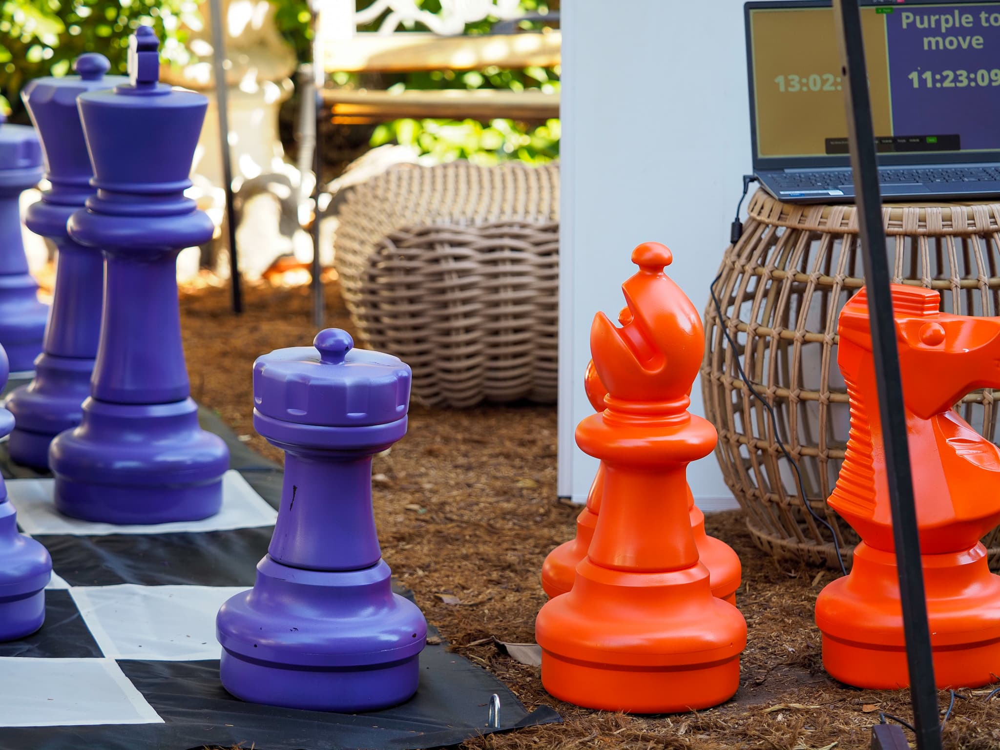 Giant purple and orange chess pieces mid-game on the Lighthaven lawn, with a laptop scoreboard reading 'Purple to move'